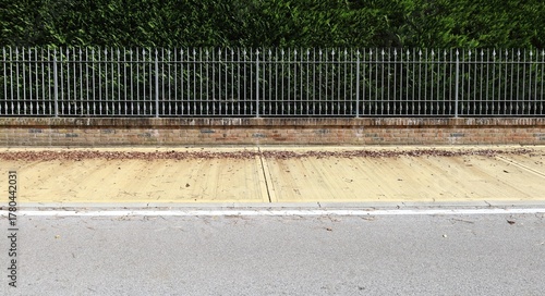 Elegant fence made of metal railing with spikes on brickwall base. High hedge on behind, yellow sidewalk and street in front. Background for copy space.