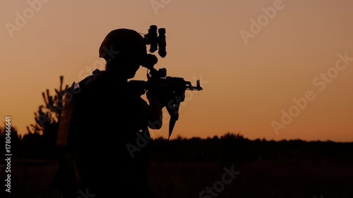 Silhouette of an armed soldier with a rifle and night vision gear against a sunset sky. Military training and tactical operation