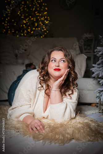 A portrait of a beautiful,chubby  woman with a sweet smile against a New Year's backdrop. A buxom woman in a white knitted sweater with an open neckline poses against a backdrop of Christmas lights.