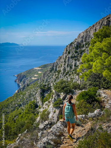 Fit female hiker with backpack walks along narrow cliffside trail with stunning view of blue sea below. Authentic moment of summer adventure and outdoor exploration along the sunny island of Hvar.