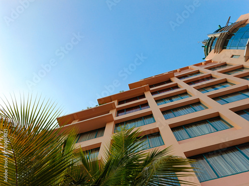 Low-angle view of a modern hotel building with palm leaves in the foreground and a bright blue sky above. Perfect for illustrating themes of travel, vacation, tourism, tropical