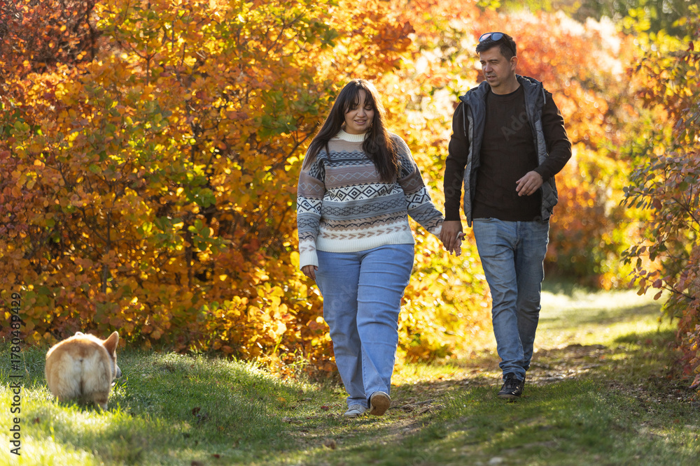 Fototapeta premium A young couple in love walks through the park with their pet. The lovers enjoy the golden autumn scenery.