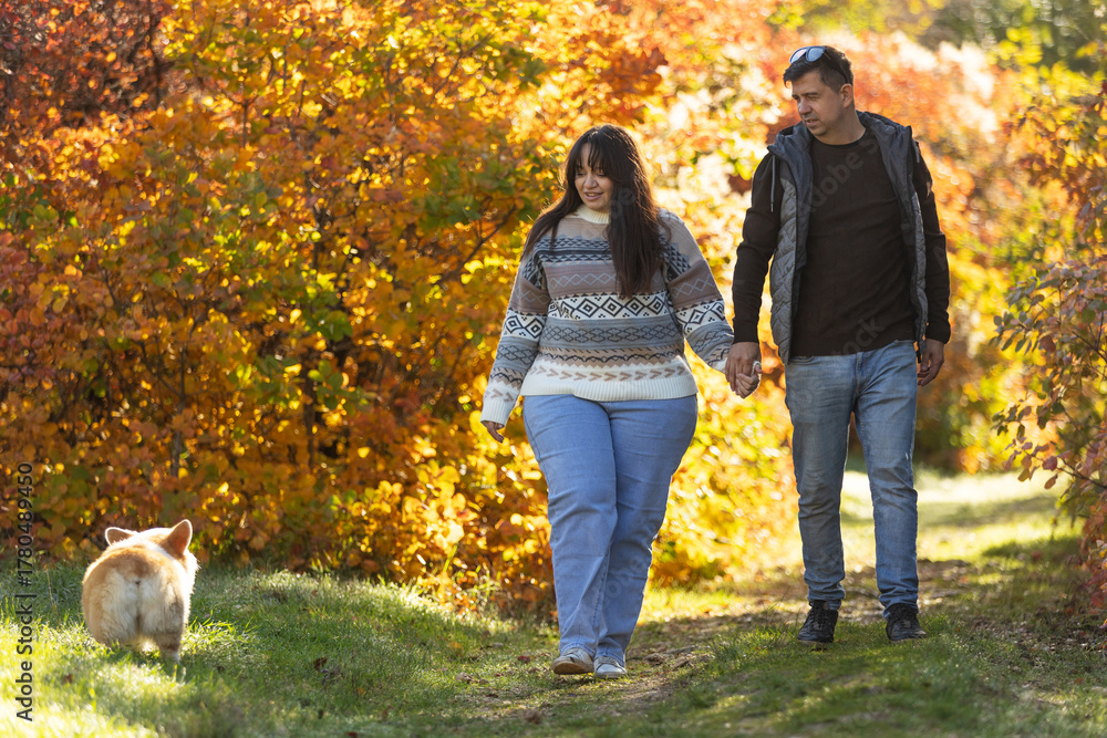 Fototapeta premium A young couple in love walks through the park with their pet. The lovers enjoy the golden autumn scenery.