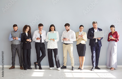 Diverse group of business professionals standing together on a gray background, working online using phones and laptops as a team. Internet technology, and collaboration in an office setting.