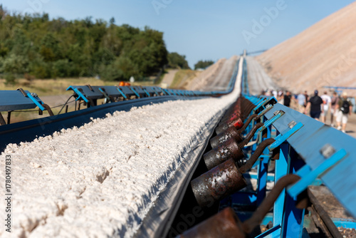 Obraz na plátně Long blue conveyor belt transports white potash ore material toward massive potassium mining dump sunlight