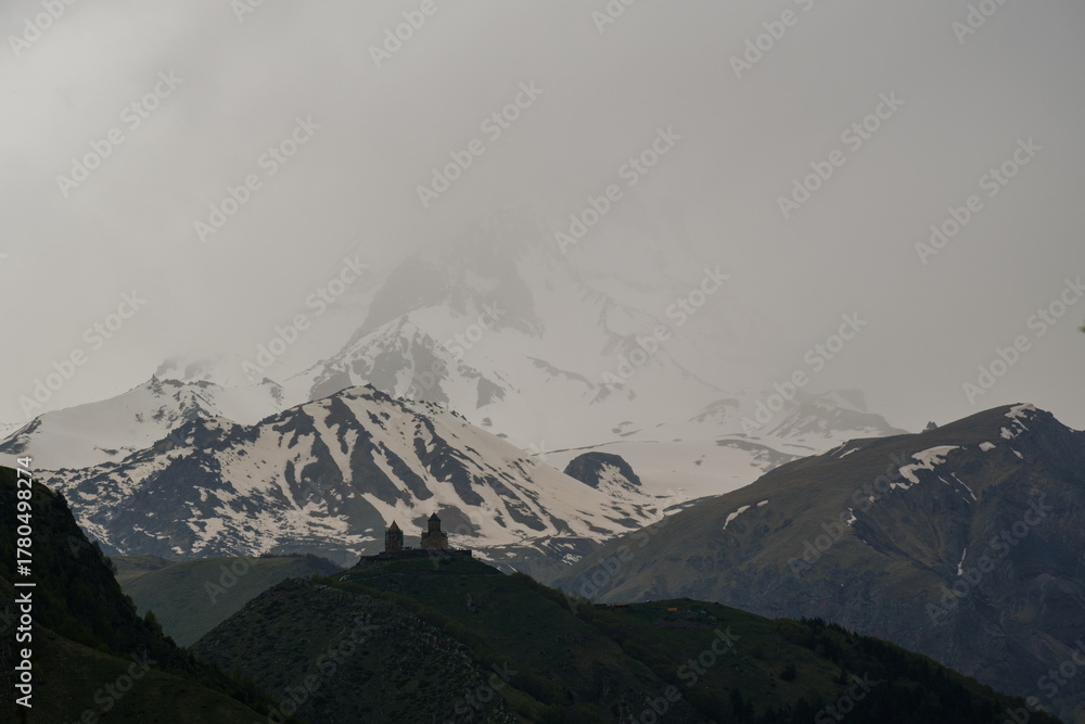 Obraz premium Snowy Mountain Kazbek with ancient Gergeti Trinity church Tsminda Sameba On a Green Hilltop Under Cloudy Skies