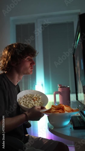 Vertical screen young man leaning forward with popcorn and chips reacting emotionally to what he sees on the glowing monitor showing deep engagement excitement and focused attention during a late even