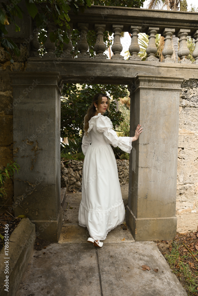 Fototapeta premium Full length female figure of beautiful blonde model wearing white fantasy wedding gown. walking through doorway of historical fairytale castle location 