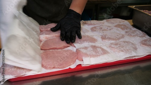 A gloved hand presses paper towel over fresh pork cutlets on a red cutting board atop stainless steel, emphasizing moisture control and hygienic meat handling