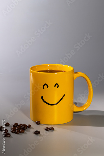 A cheerful yellow mug featuring a smiley face sits on a clean surface with coffee inside. Coffee beans are scattered nearby, enhancing the positive morning vibe.