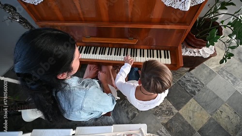 A child explores the piano keys while sitting beside their mother, both immersed in the music. Sunlight pours into the room, creating a warm and inviting atmosphere.