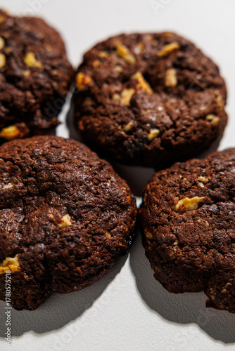 Four rich, dark chocolate cookies with visible nut chunks are arranged closely on a white background, emphasizing texture and ingredients. No branding is visible