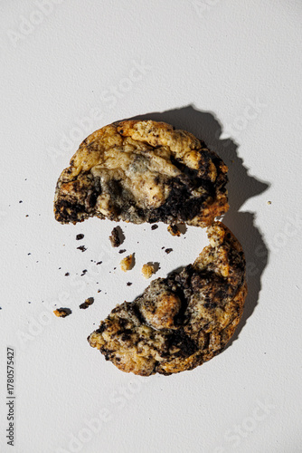 A single cookies and cream cookie is split in half on a white surface, surrounded by scattered crumbs and casting a soft shadow with visible texture and copy space