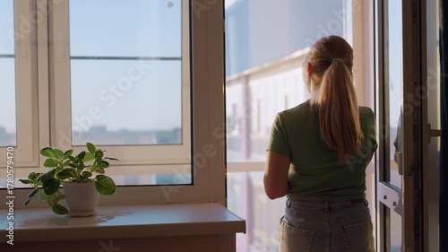 Lady with ponytail wearing green shirt walks toward balcony while on call, standing near bright window with potted green plant on windowsill, sunlight casting warm glow across home