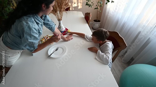 A caring mother interacts with her son at the kitchen table, playfully offering him a bite of a nutritious meal. Sunlight streams through the window, creating a cozy atmosphere.