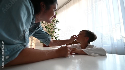 A mother interacts joyfully with her child at a bright table, sharing laughter and playful gestures as sunlight filters through the curtains, creating a warm ambiance.