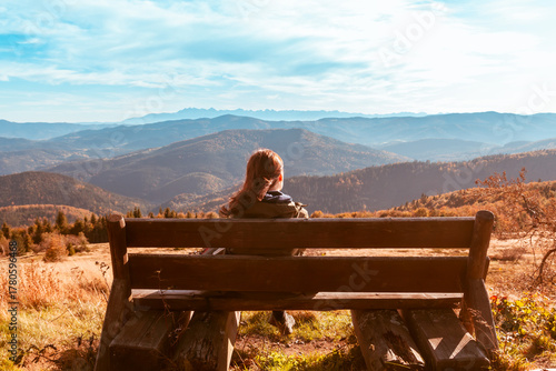 Fototapeta Naklejka Na Ścianę i Meble -  A lonely girl sits on a bench and enjoys the stunning view of the autumn mountains