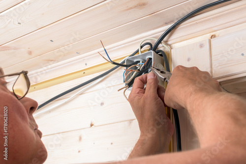 Man with glasses attentively connects electrical wires in junction box, performing installation of home wiring in wooden interior.