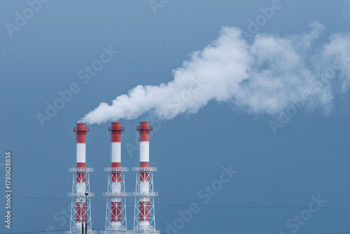 Three industrial chimneys release thick white smoke into the blue sky, symbolizing air pollution and environmental impact.