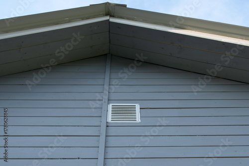 Blue wooden gable with white ventilation grille under pale sky, horizontal siding and roofline that emphasizes home airflow.