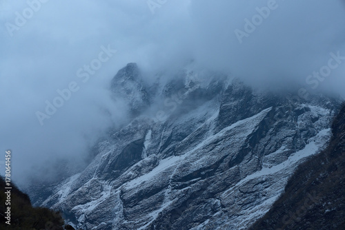 Mountain peaks are covered in snow and partially obscured by thick, low-hanging clouds. This is along the trekking route to Annapurna Base Camp in the Himalayas, Nepal