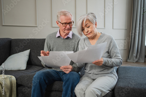 Sad tired disappointed middle aged senior couple sit with paper document. Unhappy older mature man woman reading paper bill managing bank finances calculating taxes planning loan debt pension payment