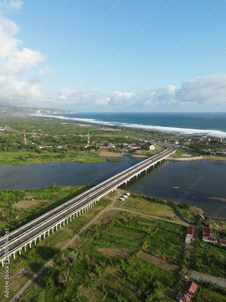 Fototapeta premium Special Region of Yogyakarta, Indonesia - October 15, 2025 : Aerial View Of Coastal Town With Bridge Over Water And Green Fields