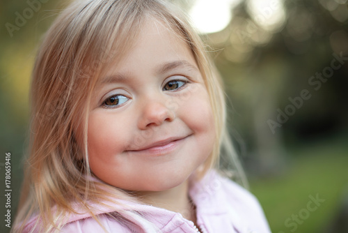 Close-up portrait of a smiling little girl outdoors. Cute child with blond hair and brown eyes looking sideways with a gentle expression. Natural light, soft focus background, warm tones. 