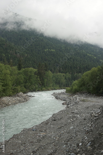 A stormy mountain river flows between rocks, boulders and cliffs in Arkhyz against a backdrop of mountains and dense forests, hidden by clouds and thick fog.