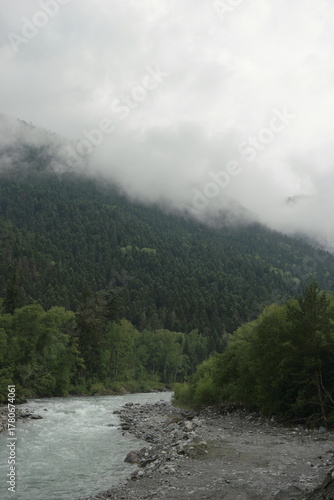 A stormy mountain river flows between rocks, boulders and cliffs in Arkhyz against a backdrop of mountains and dense forests, hidden by clouds and thick fog.