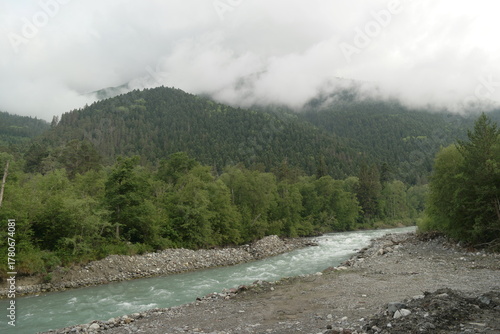 A stormy mountain river flows between rocks, boulders and cliffs in Arkhyz against a backdrop of mountains and dense forests, hidden by clouds and thick fog.