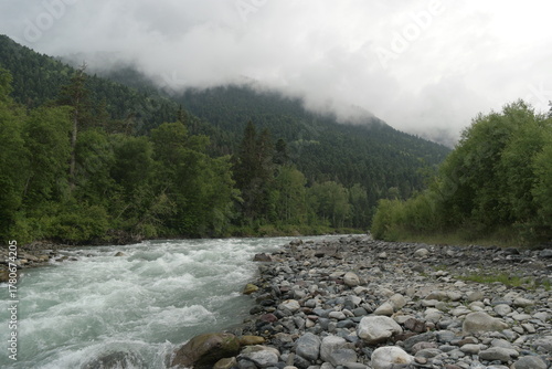 A stormy mountain river flows between rocks, boulders and cliffs in Arkhyz against a backdrop of mountains and dense forests, hidden by clouds and thick fog.