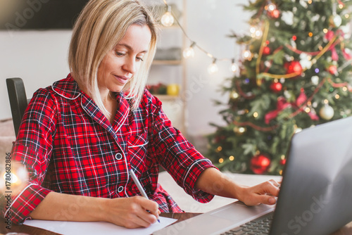 Attractive Middle aged woman works at home remotely with laptop on the background of Christmas tree