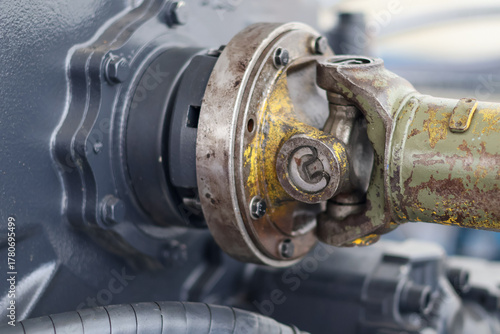 Close-up of a greasy, worn universal joint and driveshaft coupling on a heavy machinery transmission, showing rust, wear, and an attached hydraulic hose.