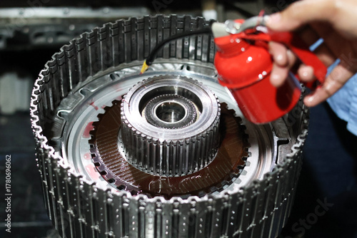 Technician applying oil from a small red pump dispenser onto the clutch pack and metallic gear components during the assembly of an automatic transmission.