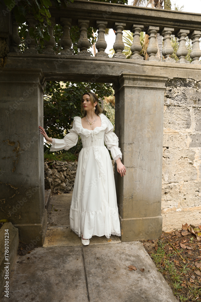 Fototapeta premium Full length female figure of beautiful blonde model wearing white fantasy wedding gown. walking through doorway of historical fairytale castle location