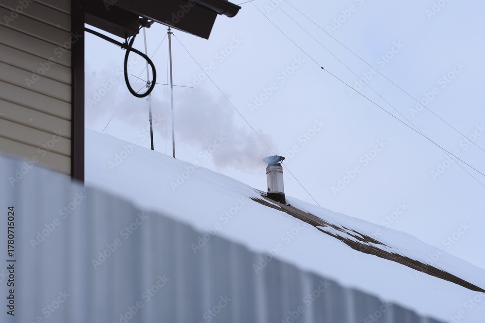 Fototapeta premium A chimney on a snow-covered rooftop releases smoke into the cold sky on a winter day.