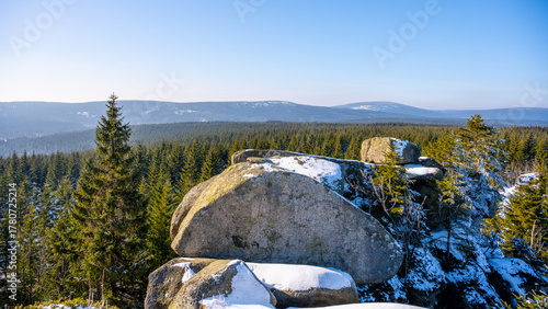 Pytlacke Rocks stand proudly amidst a snowy landscape in the Jizera Mountains. A clear morning sun illuminates the charming Jizerka village and surrounding forest.