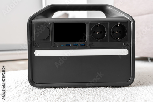 A modern portable power station sits indoors on a carpeted floor showcasing multiple outlets and USB ports for home or travel backup electricity needs.