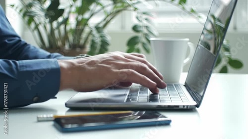 tight close-up video of male hands in blue shirt, wristwatch, typing on silver laptop on white desk with mug, phone, pencil. soft light, blurred plant. remote work productivity concept