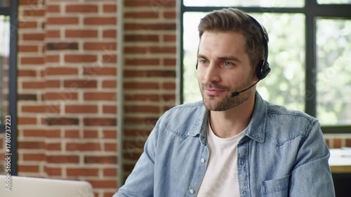 young caucasian man, 30s, in denim shirt and headset, speaks and gestures during an online video call at a wooden desk with a blurred green background, professional remote work