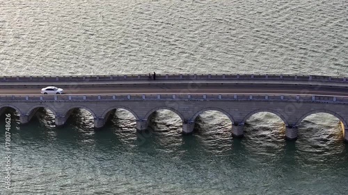 A pair of pedestrians taking a walk on the arched bridge over the lake, with a white sedan passing by