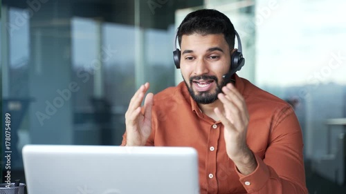Confident businessman in a headset talking on a video call using laptop sitting at workplace in office. Entrepreneur has a business meeting. Manager speaks remotely at an online conference. Close up