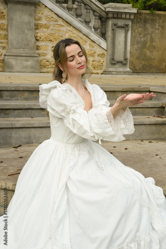 Obraz premium figure of female blonde model wearing white fantasy wedding gown. romantic sitting pose on staircase of historical fairytale castle location