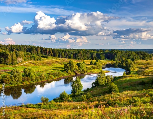 Fototapeta Naklejka Na Ścianę i Meble -  Scenic landscape captures a flowing river winding through rolling green hills and meadows beneath a partly cloudy blue sky. Lush trees border the water