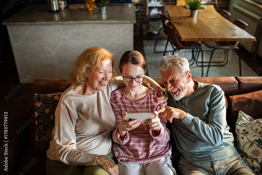 Obraz premium Senior grandparents and teen granddaughter laughing at smartphone at home