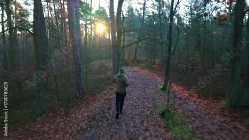 Aerial rear view of a 45-year-old man jogging through a forest on an autumn morning, illuminated by the rising sun, calmer tracking the person.