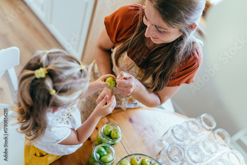 Caucasian mother teaching toddler daughter to clean greengage plums for canning at kitchen table