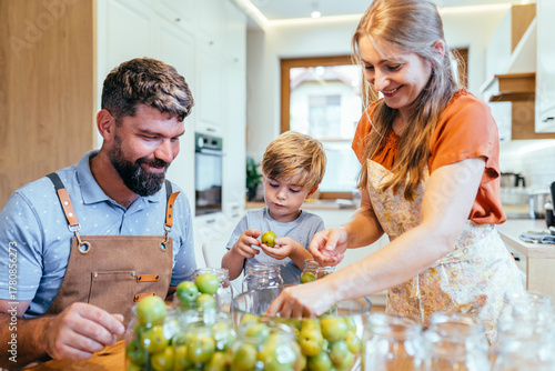 Smiling father helping son fill glass jars with greengage plums in bright home kitchen