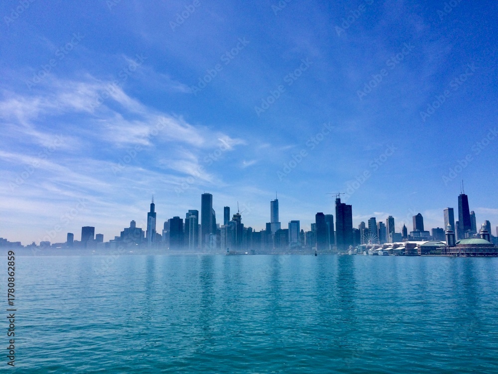 Obraz premium Dramatic panoramic view of the Chicago skyline from Lake Michigan, featuring modern skyscrapers like the John Hancock Center reflecting in serene blue water beneath a vast, clear summer sky